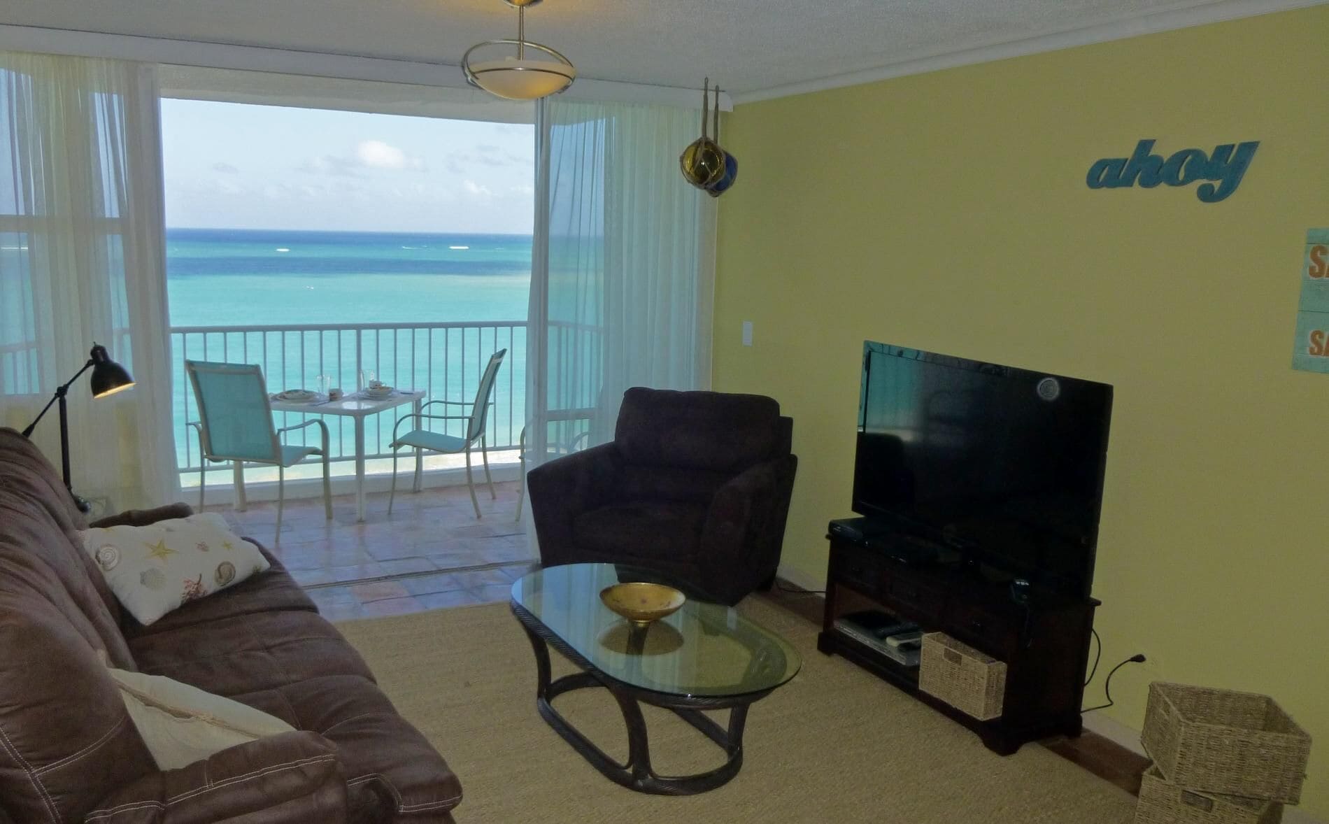 A living room with a sofa, a glass coffee table, a TV, and a balcony with a view of the ocean.