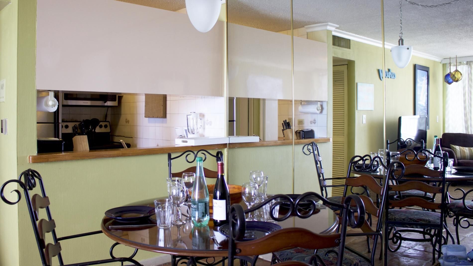 A dining area with a round table set for four, surrounded by metal chairs. A kitchen is visible in the background, and a large mirror reflects the room.