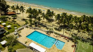 Vista aérea de un resort frente al mar, con piscina, palmeras y una playa de arena.
