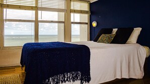 Un dormitorio con un ventanal grande, una cama con colcha blanca y azul, y vista al océano.