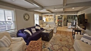 A spacious living room with a large sofa, a coffee table, and a patterned rug. There is a kitchen area in the background with modern appliances and a dining table.
