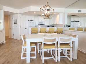 A modern kitchen with white cabinetry, a central island, and a dining area with white chairs and yellow cushions. - Cambridge 302 Beautiful Views from Large Oceanfront Balcony (Pawleys Island)