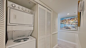 A laundry room with stacked washer and dryer units, a window with a painting, and a wooden floor. - Cambridge 302 Beautiful Views from Large Oceanfront Balcony (Pawleys Island)