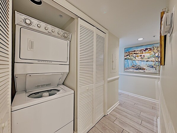 A laundry room with stacked washer and dryer units, a window with a painting, and a wooden floor. - Cambridge 302 Beautiful Views from Large Oceanfront Balcony (Pawleys Island)