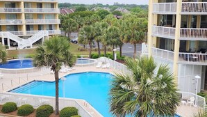 A swimming pool surrounded by palm trees and white railings, with a multi-story building in the background. - Cambridge 302 Beautiful Views from Large Oceanfront Balcony (Pawleys Island)