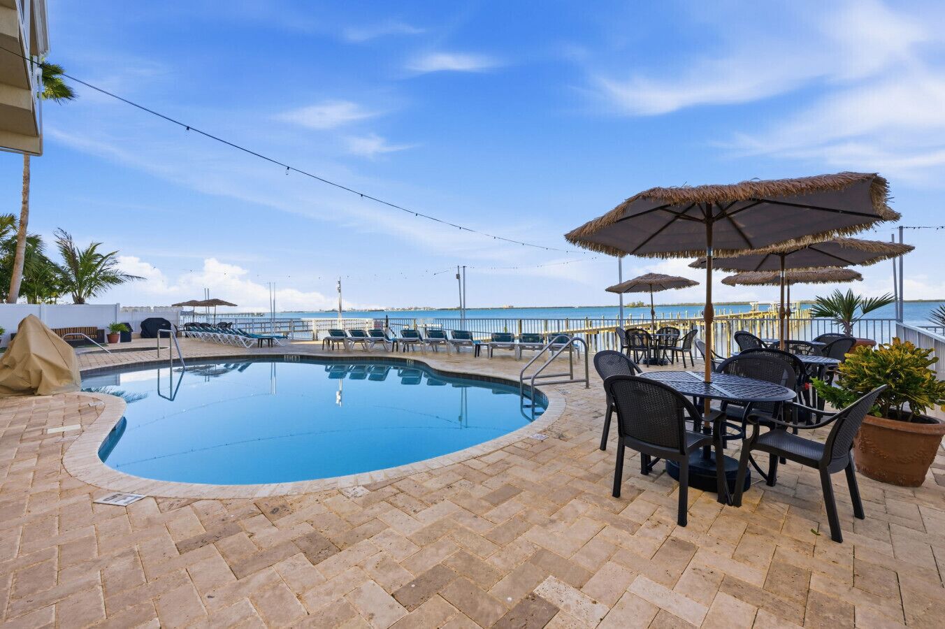 Piscina con sillones de descanso y sombrillas, rodeada de un patio de ladrillo y con vistas a un cuerpo de agua.