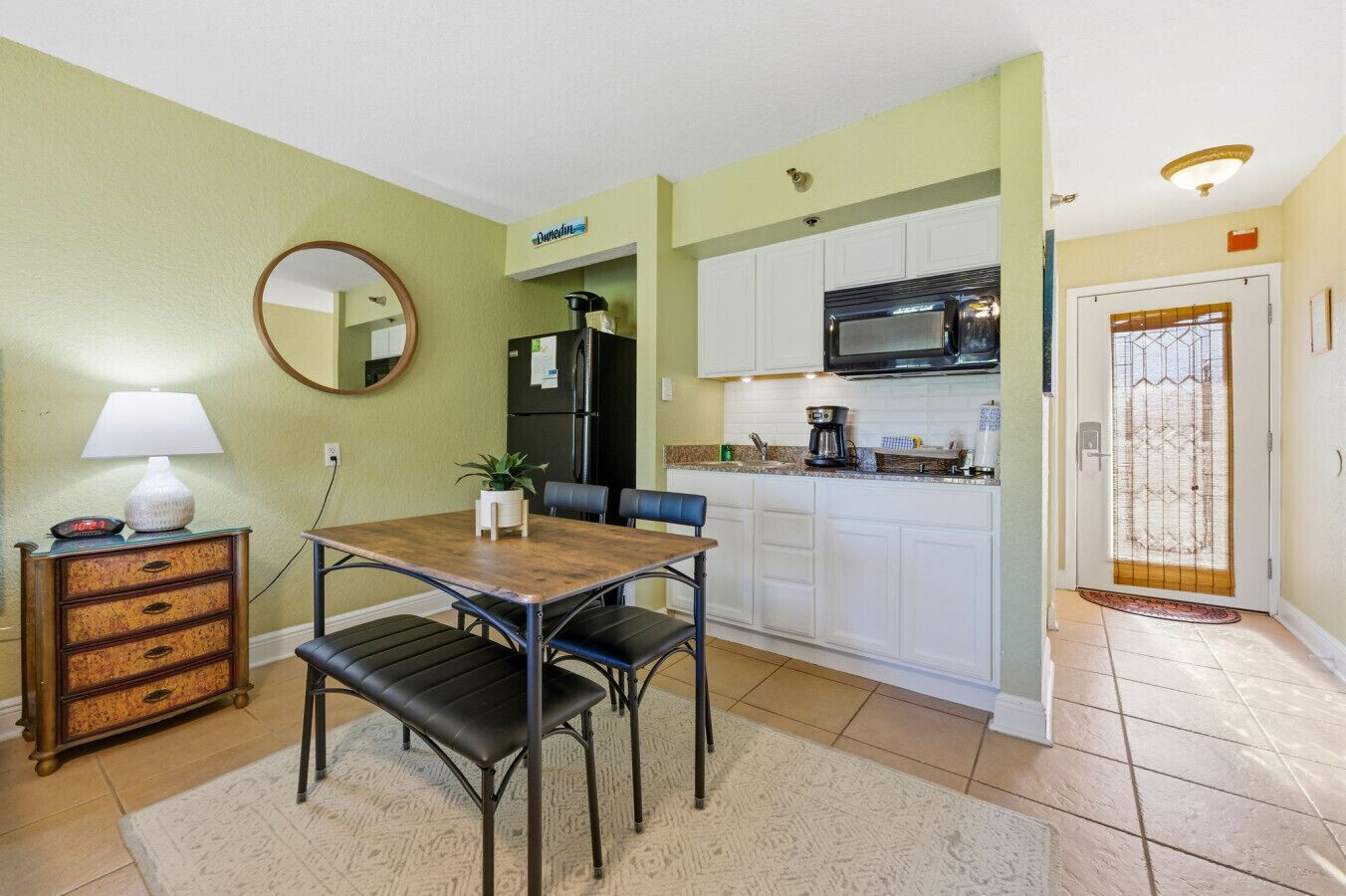 Cocina compacta con armarios blancos, un refrigerador negro, microondas y cafetera. Hay una mesa de comedor con dos sillas y una mesita auxiliar con una lámpara.