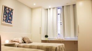 Une chambre avec deux lits, une fenêtre avec des rideaux, une petite plante sur le rebord de la fenêtre et un tableau encadré au mur.