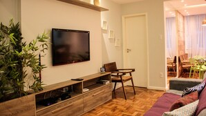 Un salon avec un t éléviseur à écran plat fixé au mur, un meuble de rangement en bois contenant des appareils électroniques, un fauteuil vert et un canapé violet.