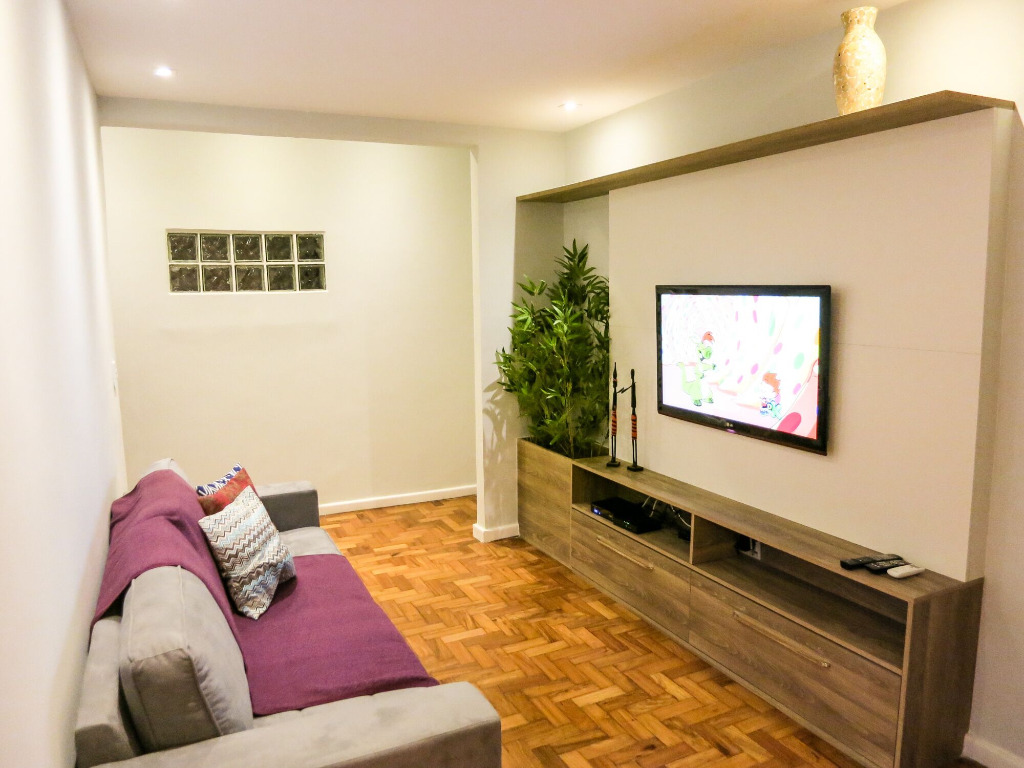 A living room with a wooden floor, a grey sofa, a television mounted on the wall, and a small plant on the side.