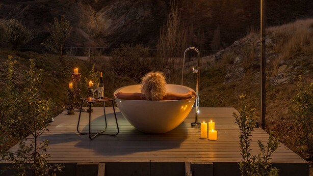 A wooden deck with a large bowl-shaped bathtub, candles, and a table with bottles and glasses.