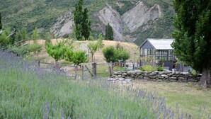 A rustic cabin with a stone wall and a metal roof, surrounded by a field of lavender and a mountainous backdrop.