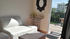 Ein Wohnzimmer mit einem weißen Sofa, einem kleinen Tisch mit Lampe und Blick auf ein Gebäude durch eine Glastür.