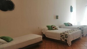 Ein Schlafsaalzimmer mit drei Einzelbetten, jedes mit einem grünen Kopfkissen und einer blumigen Bettdecke. Das Zimmer hat weiße Wände und einen gefliesten Boden.