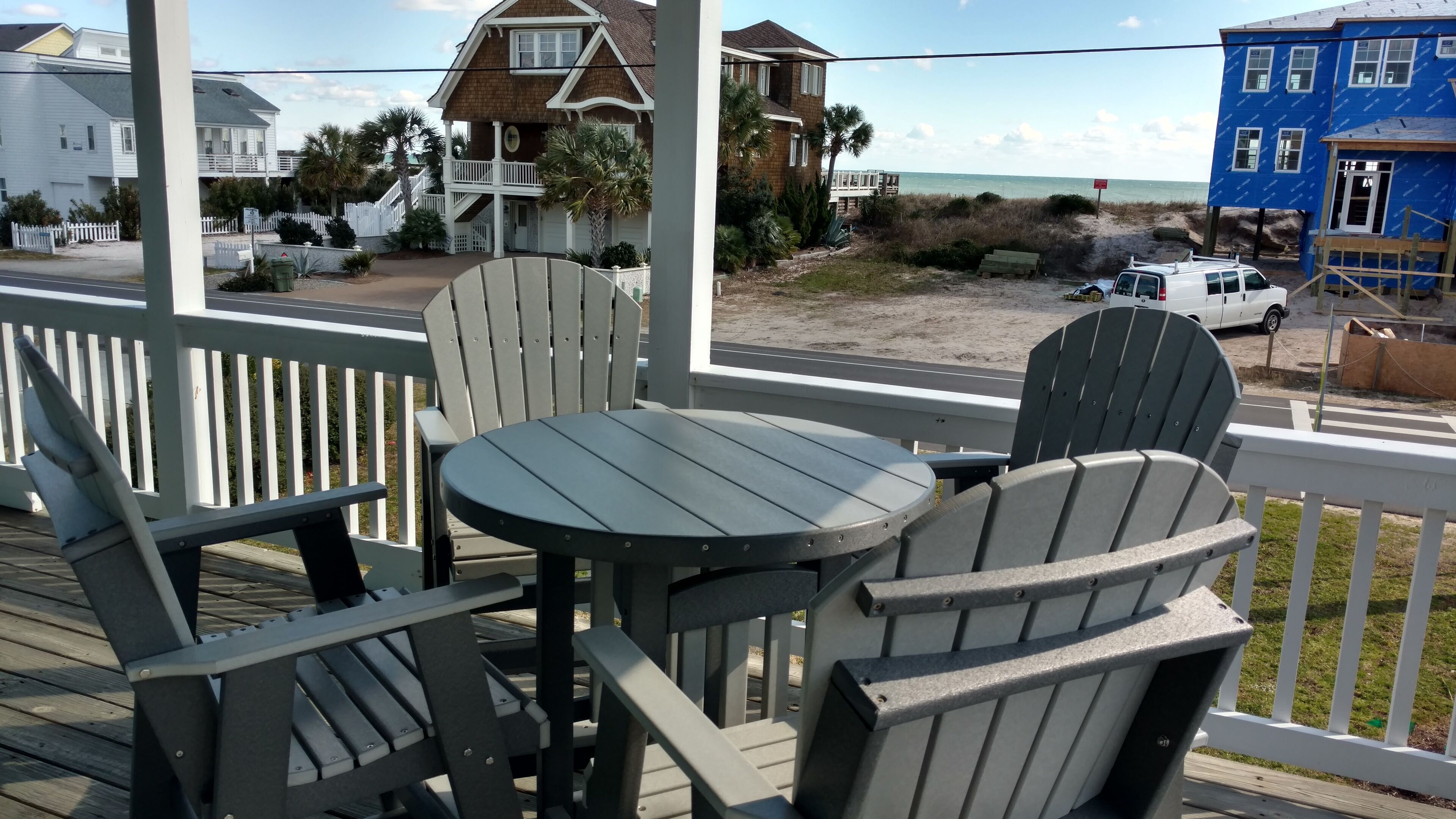Terraza de madera con sillas Adirondack y una pequeña mesa redonda, con vista a una playa y una zona residencial con casas y un edificio azul.