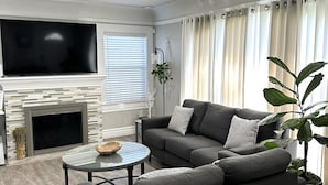 Un salon moderne avec un canapé gris, un téléviseur à écran plat, une cheminée et une grande fenêtre avec des rideaux.