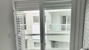 Une chambre à coucher avec un lit, une table de chevet, un vase de fleurs et une vue sur un immeuble d’habitation, visible à travers une porte coulissante.