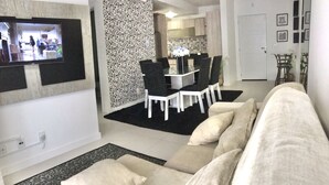 Un salon avec un canapé beige, un coin repas avec une table et des chaises en verre, et une cuisine à l’arrière-plan.
