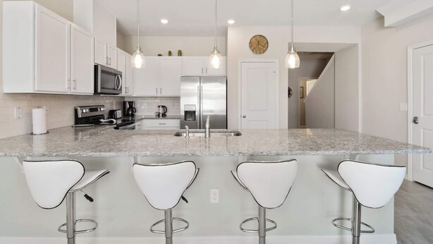 A modern kitchen with white cabinets, stainless steel appliances, and a granite countertop. There are four white bar stools and pendant lights above the island. - Family 4Bd Home Game Room Resort Near Disney 5147 (Kissimmee)