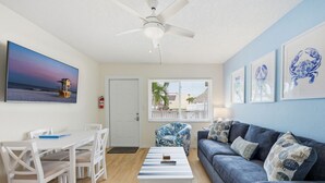 Una sala de estar con un ventilador de techo blanco, un sofá azul, una mesa de comedor blanca con sillas y un cuadro grande enmarcados de una escena de playa.
