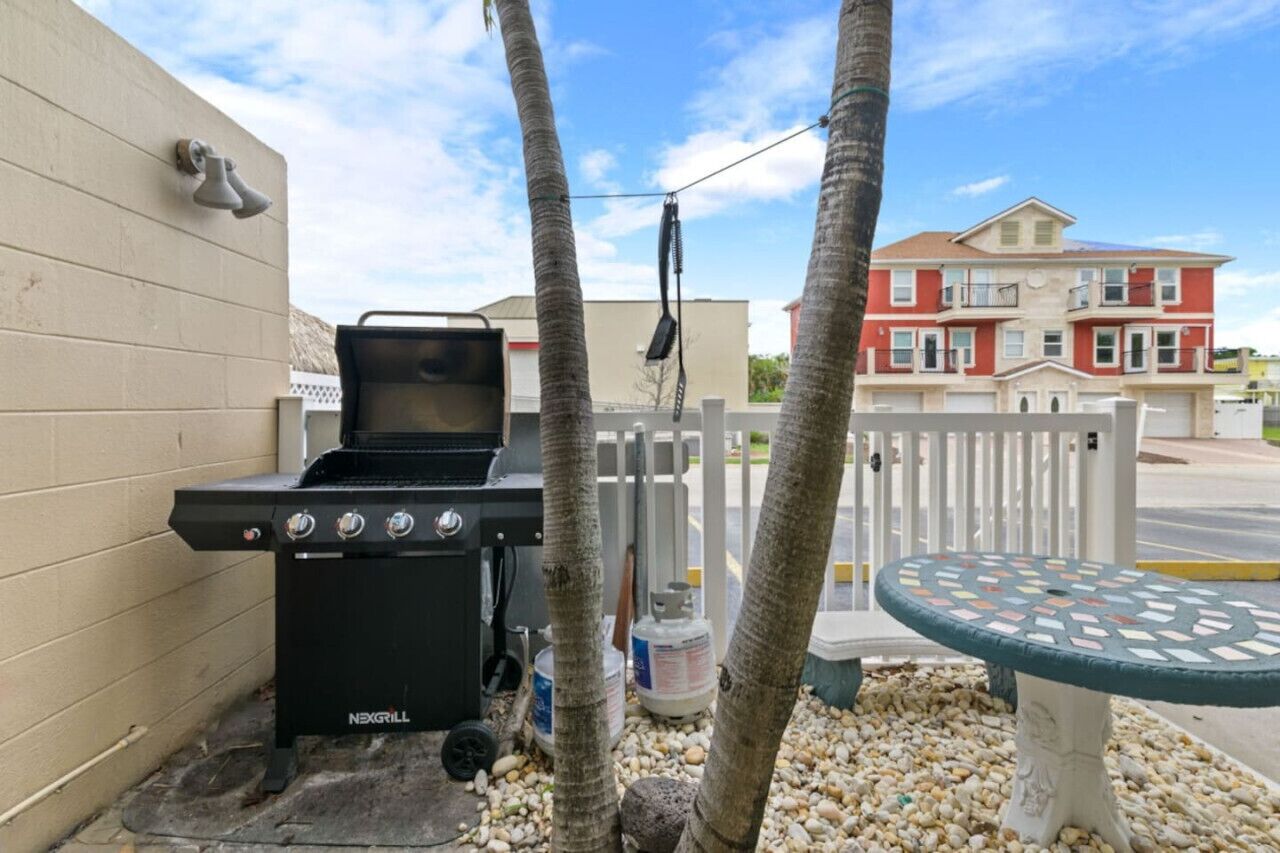A black NexGrill barbecue grill with propane tanks, a white fence, and a red building in the background.