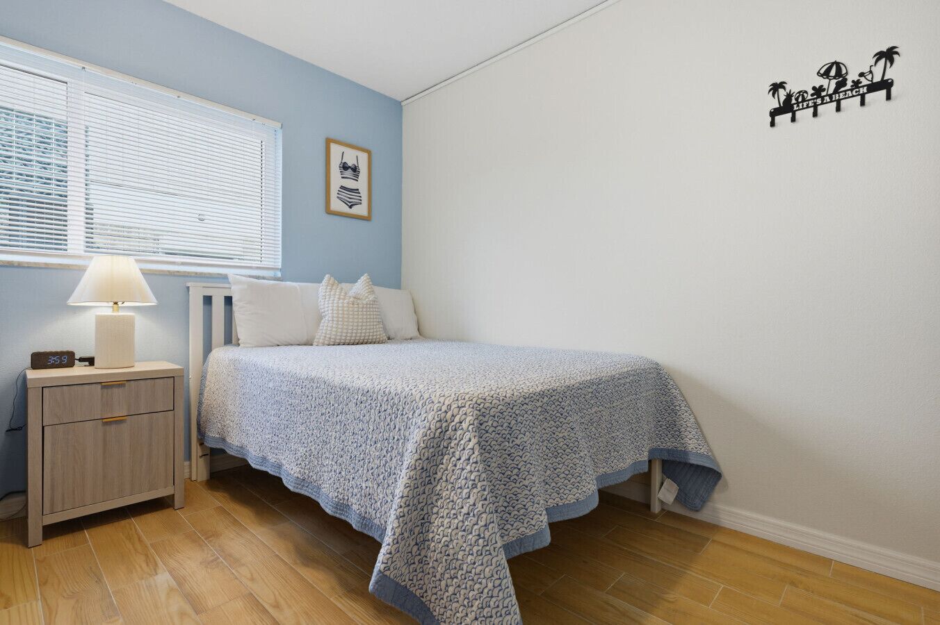 Un dormitorio con cama, mesita de noche, lámpara, ventana con persianas, un adorno en la pared y una estantería.