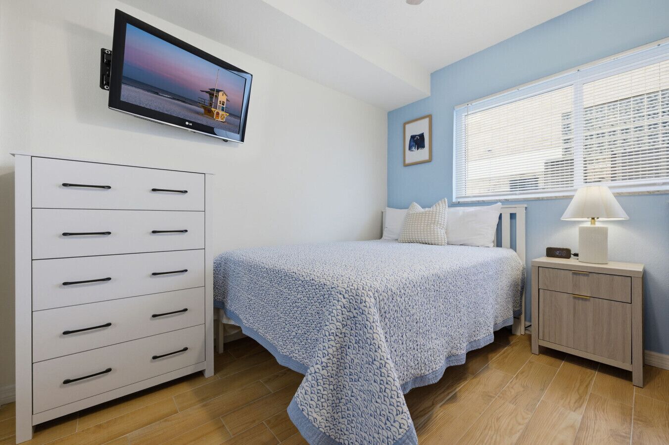 Une chambre à coucher comprenant un lit, une commode, un téléviseur, une fenêtre avec des stores et une table de chevet avec une lampe.