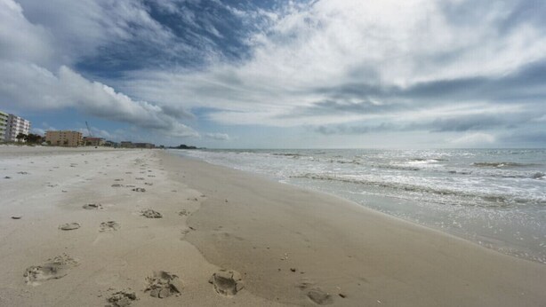Una playa con huellas que conducen al agua, cielo nublado y edificios al fondo.