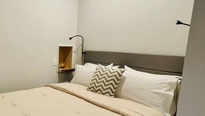 A neatly made bed with a beige comforter and pillows, a bedside table with a lamp, and a wall-mounted shelf. - Launchpad~3 Beds,  Rain Shower, Top Golf NearBy (Thornton)