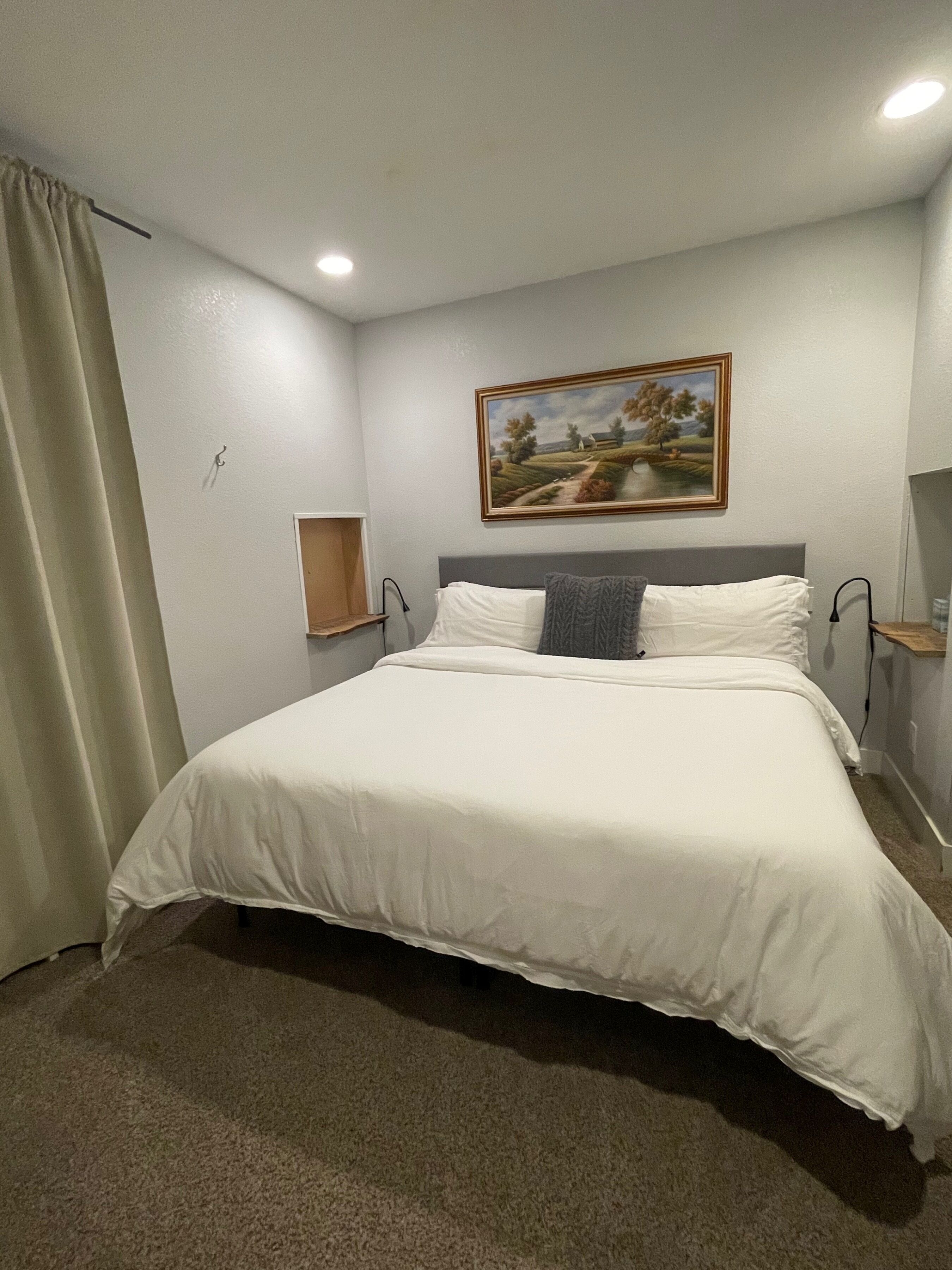 A neatly made bed in a hotel room with a painting on the wall, a small wooden shelf, and a curtain on the left.