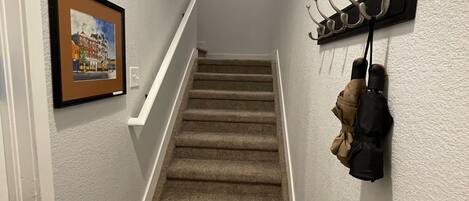 Un escalier recouvert de moquette avec une rampe blanche, un porte-manteau avec des vestes et un tableau encadré accroché au mur.