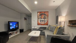 A modern living room with a flat-screen TV, a grey sofa, a white coffee table, and a lamp. - Launchpad~3 Beds,  Rain Shower, Top Golf NearBy (Thornton)