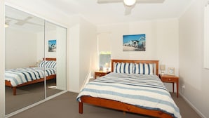 A bedroom with a large mirror, a bed with striped bedding, a nightstand, a lamp, and a framed picture on the wall. - Toorumbee 4/38 - Three Bedroom Townhouse Pet Friendly on Request* (Mooloolaba)
