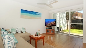 A living room with a sofa, a coffee table, a television, and a large window with a view of a backyard. - Toorumbee 4/38 - Three Bedroom Townhouse Pet Friendly on Request* (Mooloolaba)