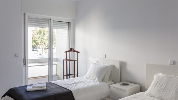 A modern bedroom with two beds, a nightstand, a wooden hanger, and a sliding glass door to the outside.