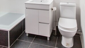 A modern bathroom with a white sink, mirror, and toilet. There is a towel rack with two towels, a glass shower enclosure, and a white storage shelf.