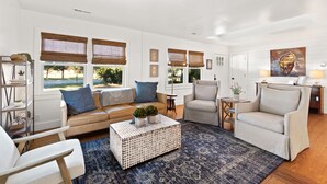 A modern living room with a beige sofa, a white coffee table, and a blue rug. There are two windows with bamboo blinds, a painting on the wall, and a small side table with a lamp.