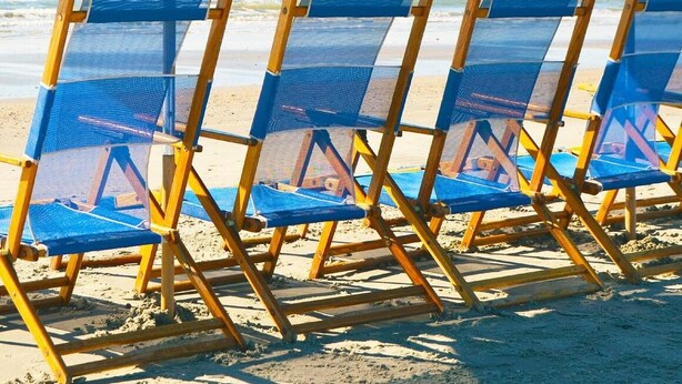 A row of blue beach chairs with yellow frames, arranged on a sandy beach with a clear view of the ocean in the background.