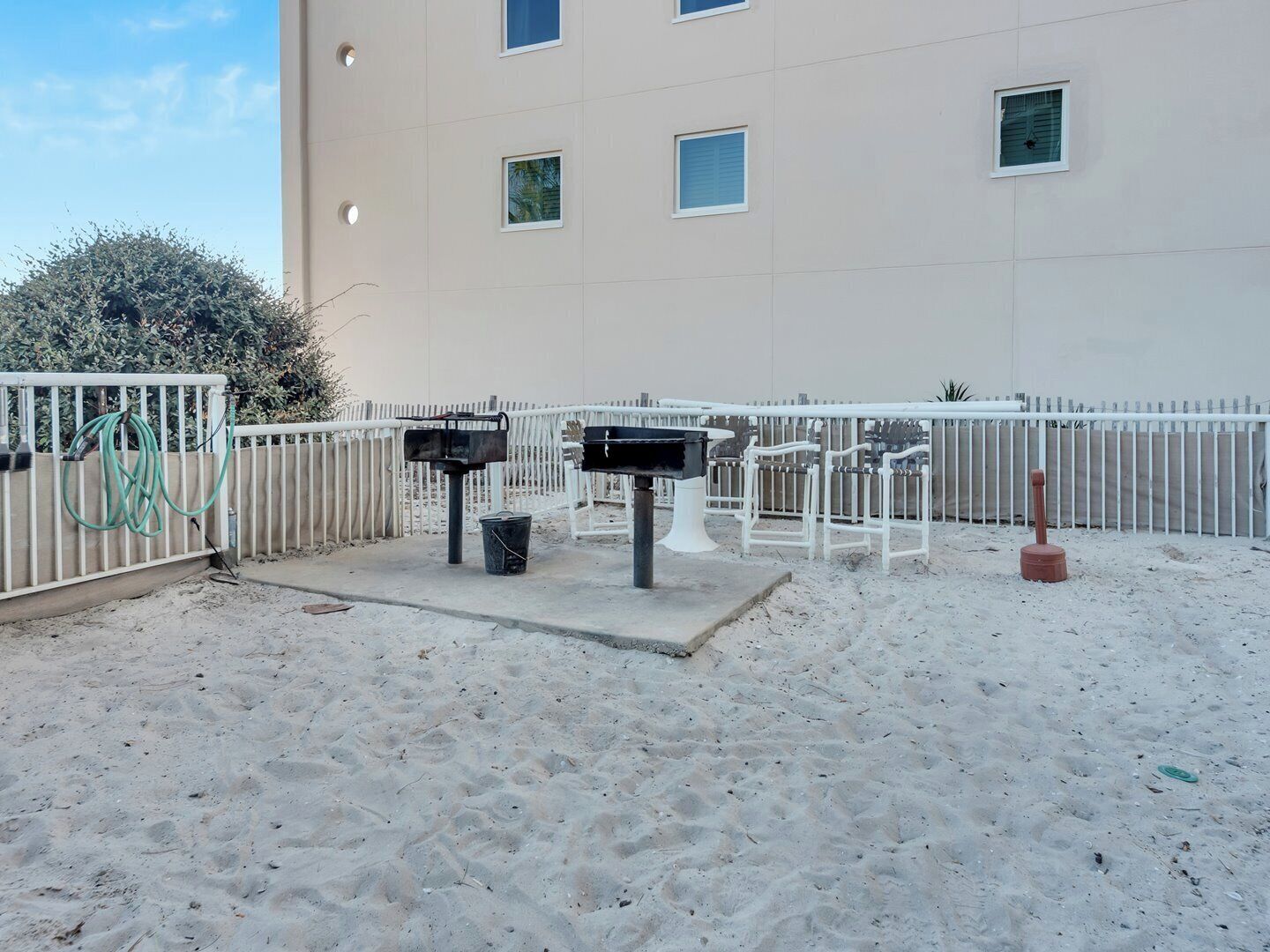 A sandy outdoor area with a white fence, two barbecue grills, and a hose.