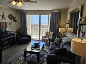 Ein Wohnzimmer mit Sofa, Sessel und Couchtisch, einem Fernseher und einem Balkon mit Aussicht.