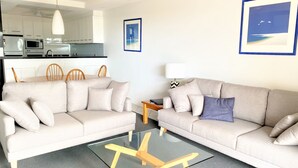 Un salon avec deux canapés beige, une table basse en verre, une table d’appoint en bois et un tableau encadré accroché au mur.