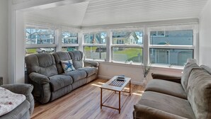 Una sala de estar con ventanales que ofrece vistas a una zona residencial, un sofá gris, una mesa de centro de madera y un banco blanco acolchado.