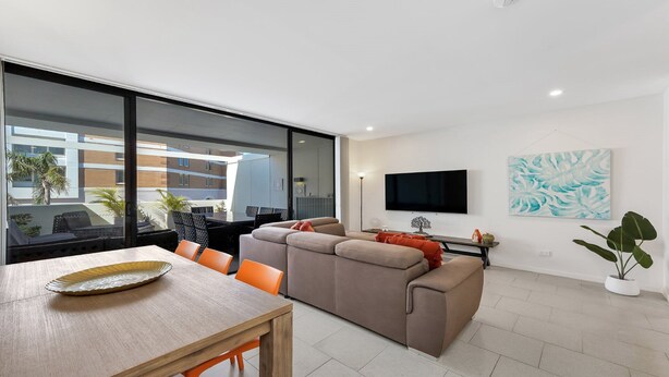 A modern living room with a sofa, dining table, and chairs, a flat-screen TV, and a large window overlooking a building. - THE HARMONY SUITE | WIFI , Short walk to beach | Accom Holidays (TERRIGAL)
