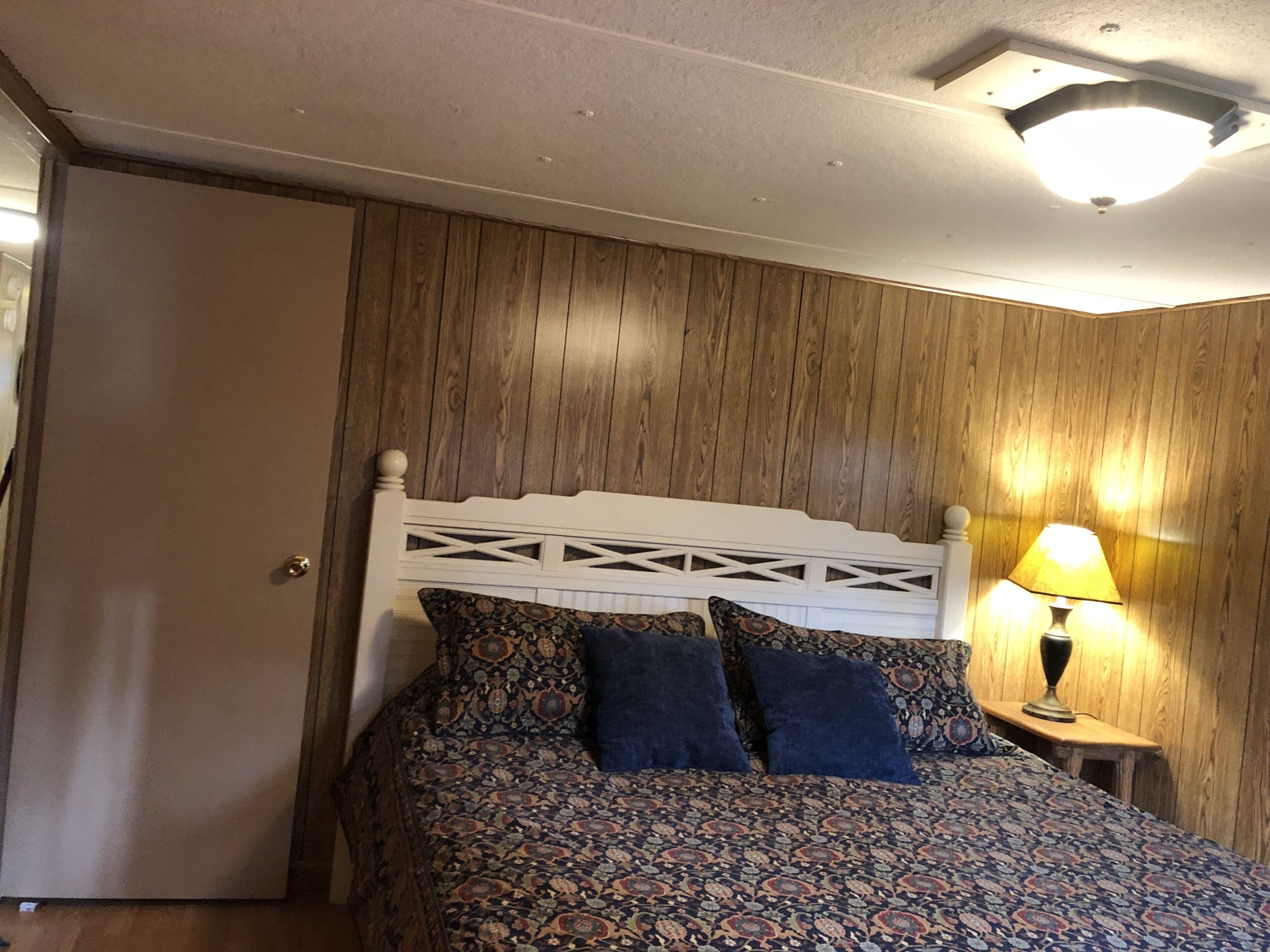 A bedroom with a wooden headboard, patterned bedspread, two dark pillows, a bedside lamp, and a door.