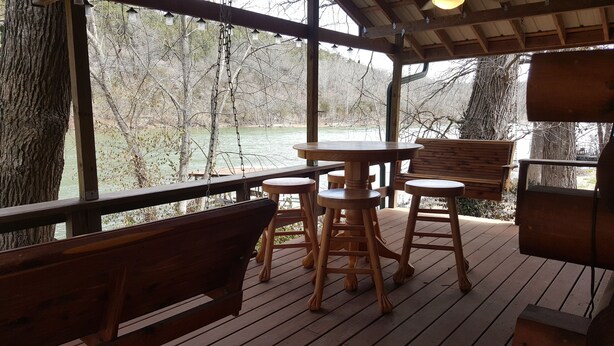 Eine Holzterrasse mit einem Tisch und zwei Barhocker, mit Blick auf ein Gewässer und Bäume.
