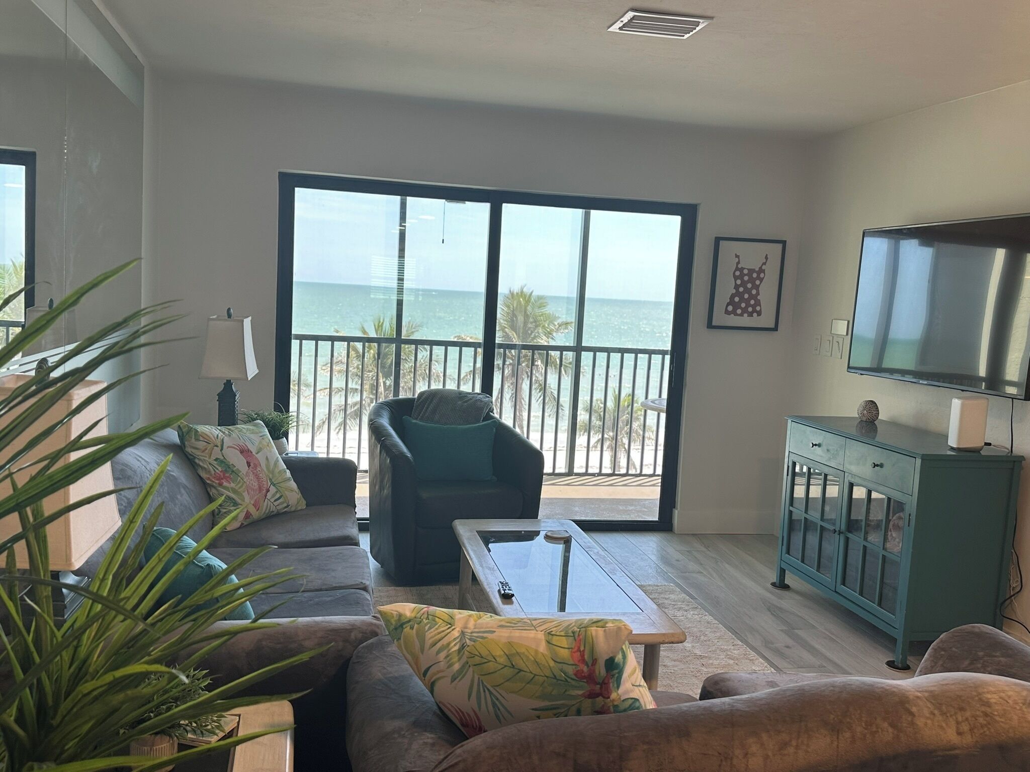 A living room with a sofa, armchair, coffee table, and a TV. There is a balcony with a view of the ocean.