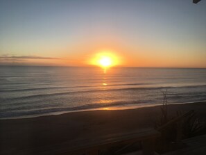Una platja amb el sol ponent-se sobre l'oceà, un pont de fusta i una barana.