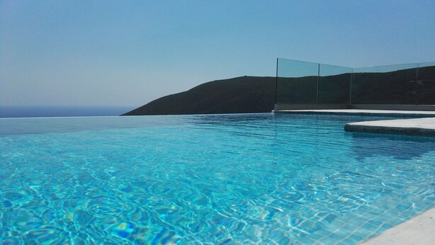 Una piscina infinita de color azul intenso con barandilla de cristal y vista al océano y una colina al fondo.