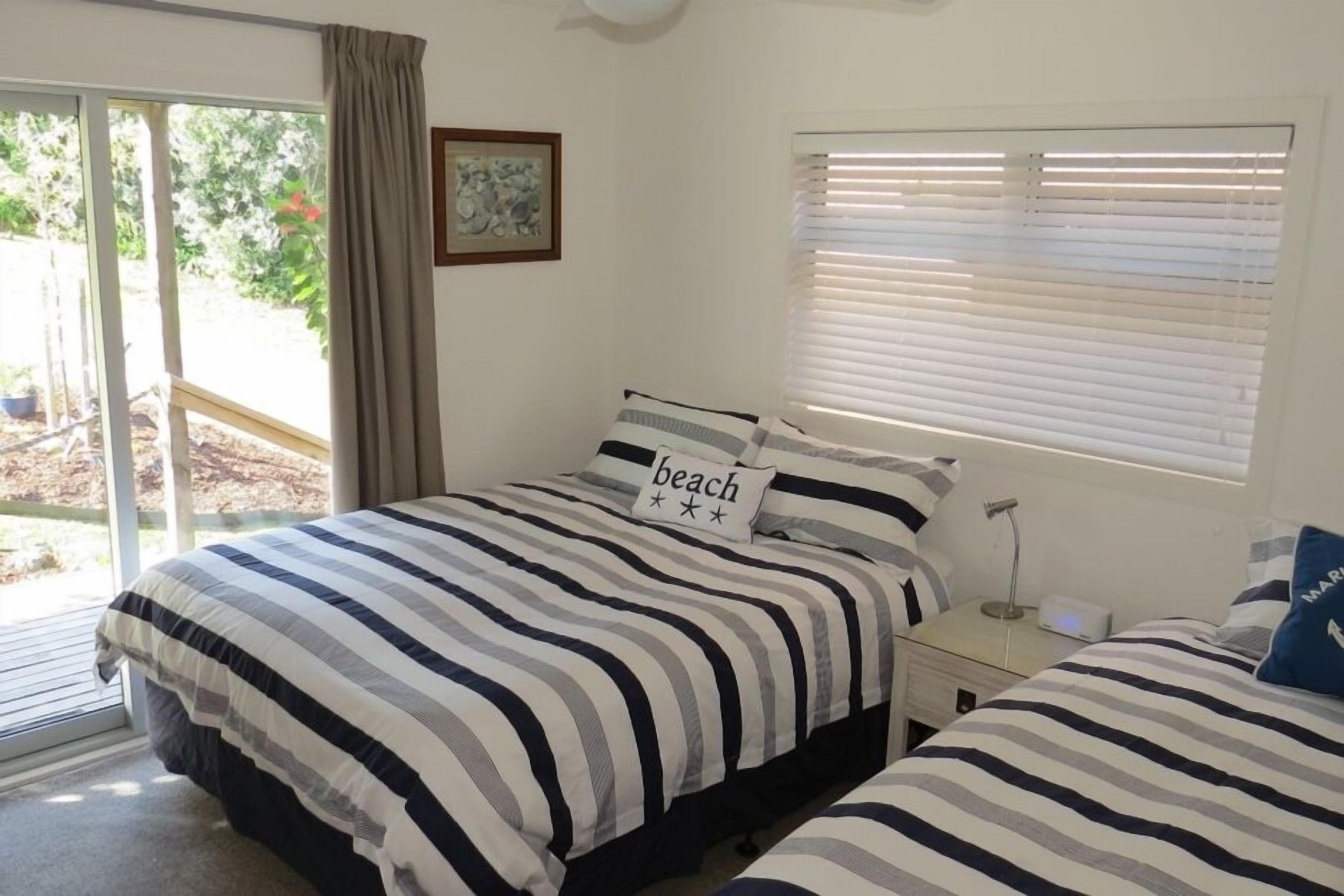 A bedroom with two beds, a window with blinds, a bedside table with a lamp, and a view of the outdoors.