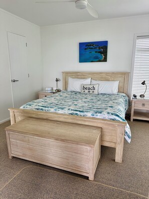 A bedroom with a wooden bed, a bench, a nightstand, a lamp, and a painting of a beach scene. - Bay of Islands Beachfront - Swim all tide, Kayaks, Sea Views - Tapeka del Mar (Tapeka Point (Russell))